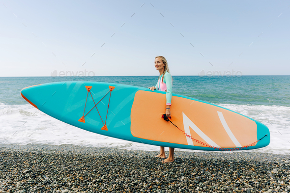 Young athletic blonde woman stands on the beach and holds SUP surf.