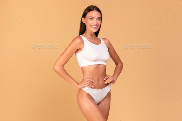 Body slimming concept. Happy lady with slim figure in white underwear smiling at camera over beige