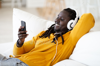 Portrait Of Smiling Black Man Wearing Headphones Relaxing With Smartphone At Home