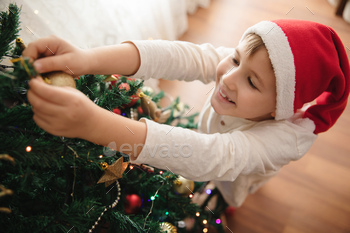 Cheerful smiling Caucasian boy wearing Santa hat decorating Christmas tree.
