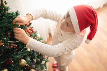 Little cute Caucasian boy in Santa's hat decorating Christmas tree at home.