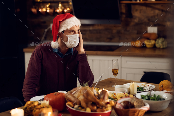 Sad man with face mask sitting alone at dining table on Christmas due to COVID-19 pandemic.