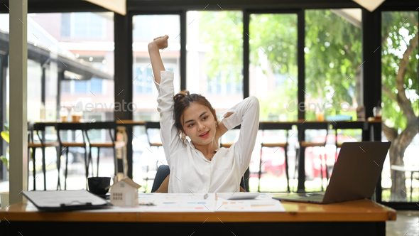 Happy young woman relaxing at workplace, reclining back in chair and stretching her arms.