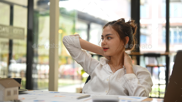 Asian female employee relaxing at work, reclining back in chair and stretching her arms.