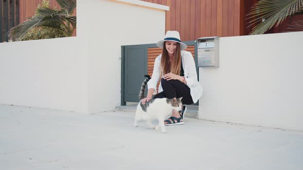 Positive Woman in Summer Hat Petting Fluffy Cat on Street alt
