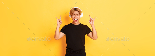 Joyful handsome asian guy enjoying listening music in headphones, showing rock-n-roll gesture