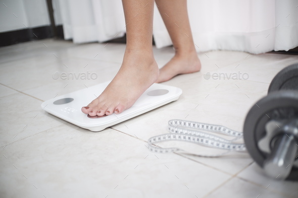Cropped image of woman feet standing on weigh scales, A tape measure and dumbbell in the foreground