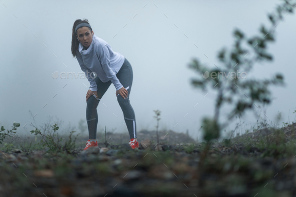 Young sportswoman bending while catching her breath after the run on misty morning.