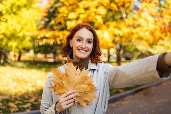Glad happy pretty european young redhead lady in raincoat hold yellow leaves, takes selfie for blog