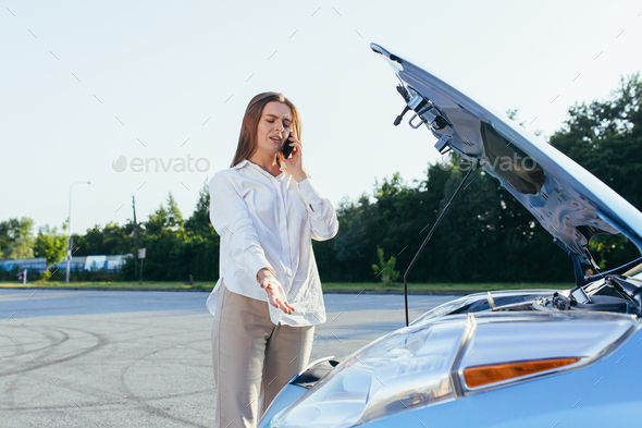 An annoyed and angry woman, near a broken car, tries to call an insurance agent and a repair crew