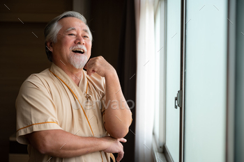 Cheerful happy senior male patient standing near window in hospital room looking out and thinking