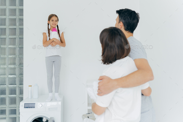 husband and wife embrace and talk to small girl standing on washer with bottle of detergent