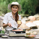 Cheerful female traveler drinking coffee and grilling bbq on a portable gas stove during camping. - PhotoDune Item for Sale