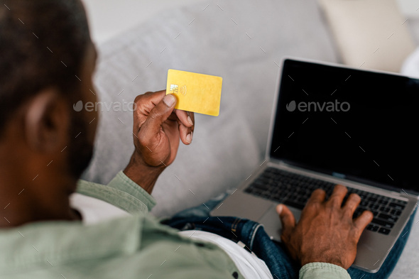 Adult african american guy in casual typing on laptop with empty screen and show credit card