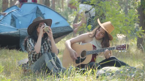 Beautiful Young Women Singing and Playing Ukulele As Men Putting Up Tent at the Background. Portrait alt
