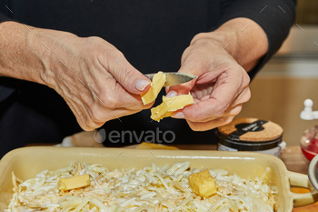 Step by step potato gratin recipe. Hands cut butter into slices of potatoes in glass baking dish