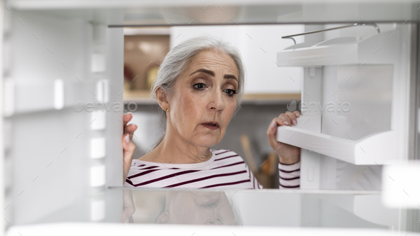 Sad Senior Woman Looking At Empty Shelves In Fridge At Home