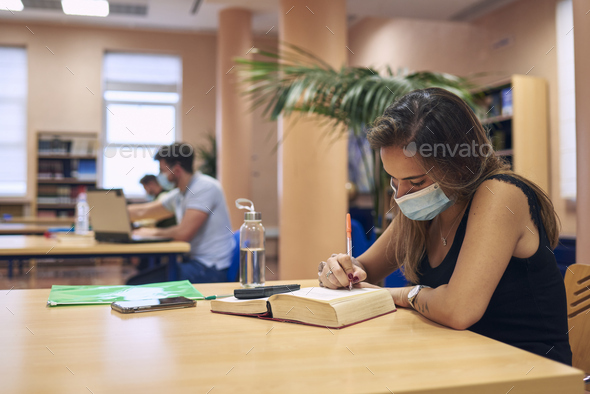 a girl in a mask studying in the library keeping a safe distance