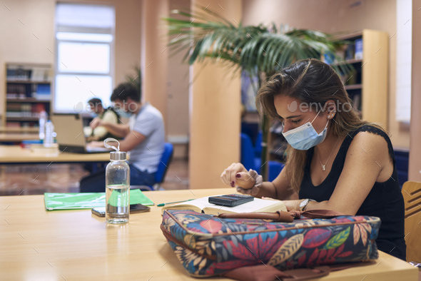 a girl in a mask studying in the library keeping a safe distance