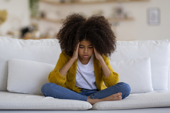 Stressed black little girl sitting on couch at home Stock Photo by