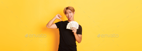Sassy handsome asian guy pointing finger at cash and looking pleased. Korean man borrowed money