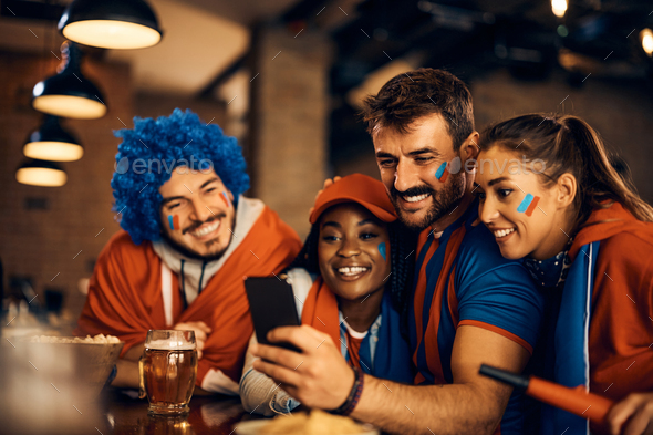 Group of happy sports fans following the match results on mobile phone in bar.