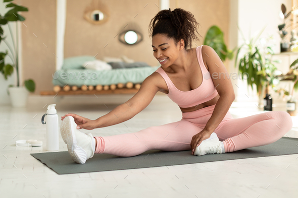 Happy black lady in sportswear exercising at home, stretching on fitness mat, touching her feet with
