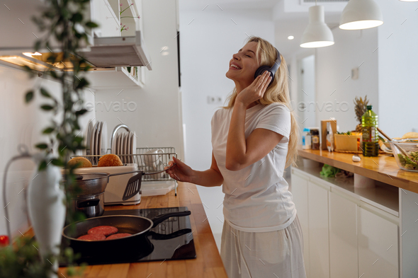 Cheerful pretty blonde woman in headphones frying pancakes and listening to music.