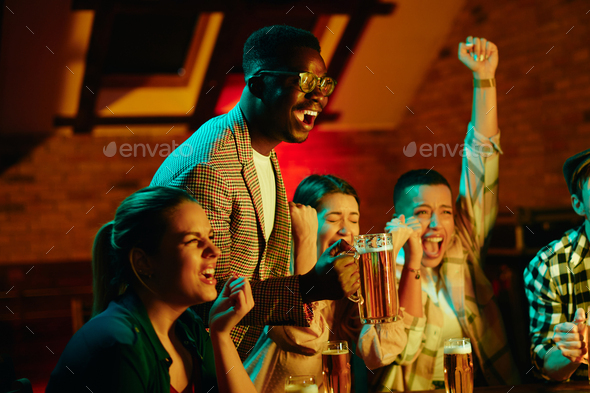 Happy African American man drinking beer and watching sports match with friends in a bar.