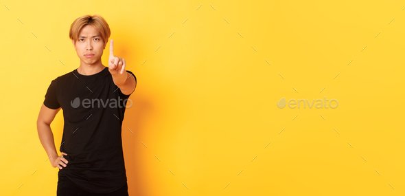 Portrait of serious-looking disappointed asian man shaking finger to scold someone, standing yellow