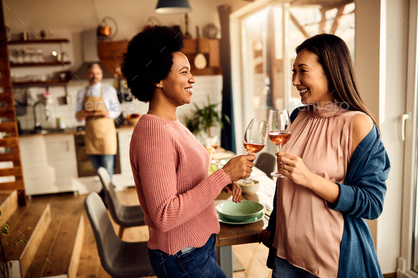 Happy black and Asian female friends toasting with wine at home.