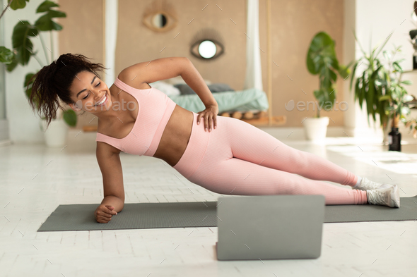 Domestic online workout. Happy black lady standing in side plank on floor at home and watching video