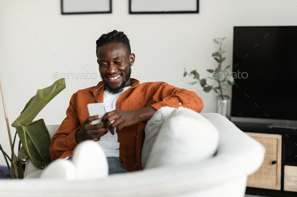 Cool gadget and app. Excited black man holding mobile phone and typing sms message, resting on sofa