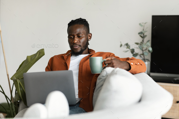 African american man working on laptop, holding cup and enjoying drinking morning coffee, sitting on - Stock Photo - Images