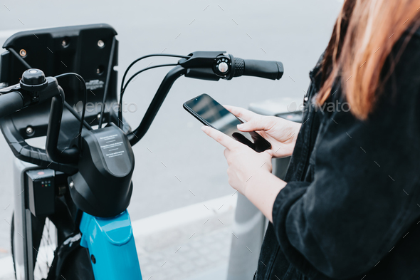 Close up of bike handlebar next to smart phone hold by young hands out of focus rental bikes. Writin