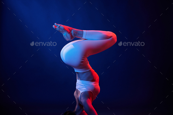 Doing head stand. Young woman in sportive clothes is in the studio with neon lights