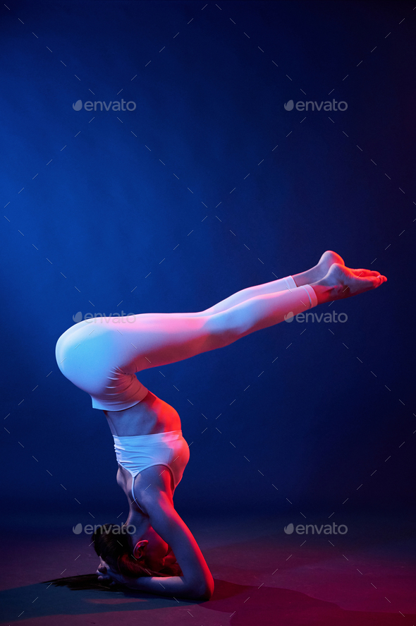 Doing head stand. Young woman in sportive clothes is in the studio with neon lights