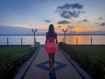 Rear view of woman in pink dress walking down an alley towards the lake at sunset