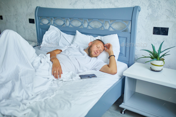 Bearded man sleeping alone on a big and cozy bed white linens with smartphone