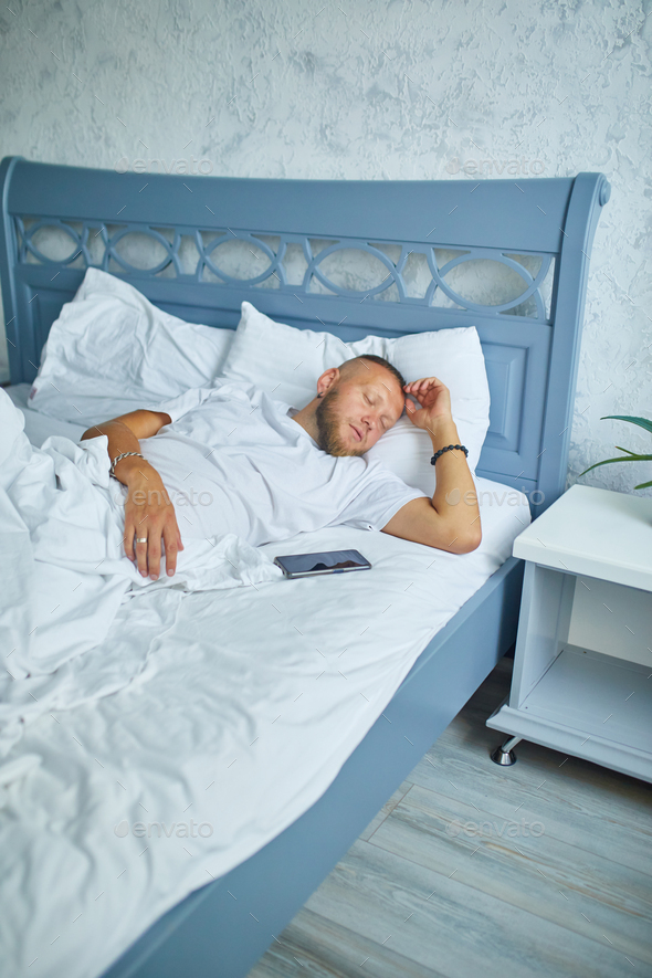 Bearded man sleeping alone on a big and cozy bed white linens with smartphone