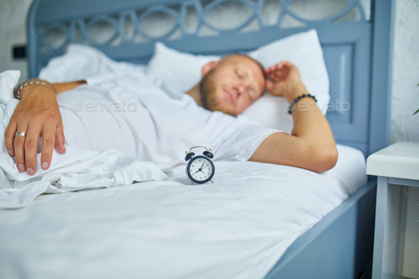 Bearded man is sleeping alone on a big and cozy bed white linens with alarm clock