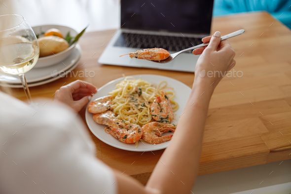 View over shoulder on plate with seafood pasta and glass of white wine. Laptop computer.
