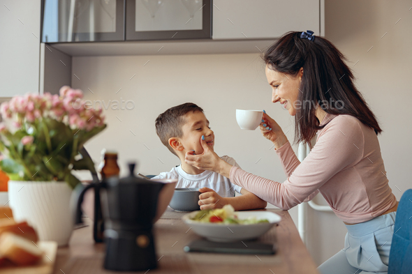 Beautiful mother drinking coffee in the morning and cleaning cheek of small son.
