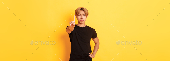 Portrait of serious-looking disappointed asian man shaking finger to scold someone, standing yellow