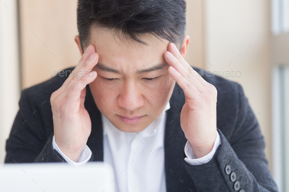 young asian man, office worker sitting at workplace holding hands massages forehead and head with