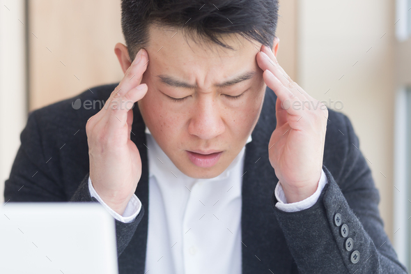 young asian man, office worker sitting at workplace holding hands massages forehead and head with