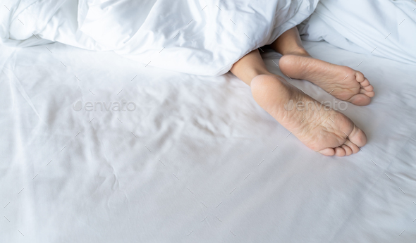 Woman barefoot on bed under white linen blanket in hotel or home bedroom. Healthy sleep