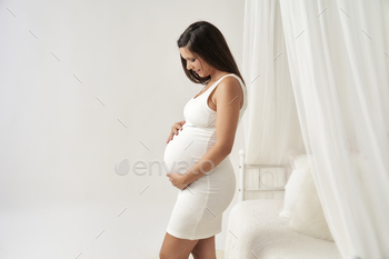 pregnant woman dressed in white touching her belly next to a white bed