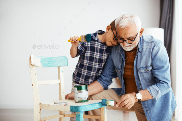 Affectionate boy embracing his grandfather while painting chair together at home.
