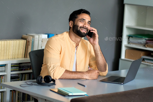 Happy indian business man sitting at work desk talking on phone making call.